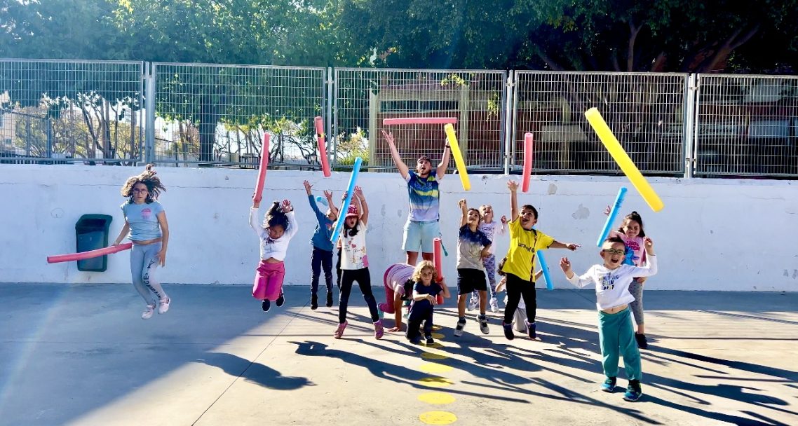 Niños saltando y celebrando al aire libre con tubos de colores en un ambiente festivo.